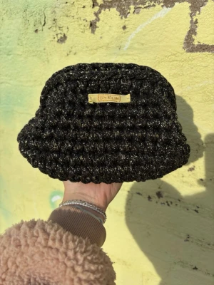 Svart glittrig clutch BY ELIN - Modell AGNES - Svart glitter - Snygg svart clutch från BY ELIN med glittrigt garn och handvirkad struktur. Väskan har en mjuk, bubblig form och en liten guldfärgad logga framtill. Perfekt accessoar för att lyfta din outfit med lite extra bling. Väskan öppnas och stängs med hjälp av en metallclips.