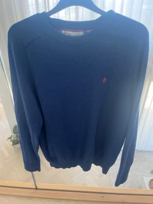 Blå stickad tröja från Penguin - Mysig blå stickad tröja från Penguin med rund halsringning och diskret röd logga broderad på bröstet. Tröjan har långa ärmar och ribbade muddar vid ärmslut och nederkant. Tillverkad i Italien. Det är en barn L så i vuxen storlek är detta en S.