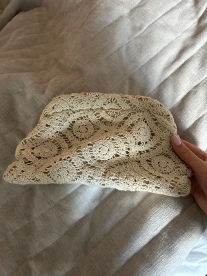 Beige virkad kuvertväska - Söt beige kuvertväska med virkat spetsmönster och mjukt innerfoder. Väskan har en klassisk rektangulär form och stängs upptill. Perfekt accessoar för att lyfta din outfit med en bohemisk touch.