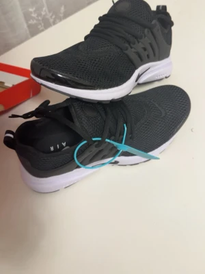 Nike Air Presto svarta sneakers - Svarta Nike Air Presto sneakers med mesh-ovandel och vita sulor. Skorna har en sportig look med plastdetaljer på sidorna och klassisk Nike-logga. Lätta och bekväma med snörning och låg profil. Perfekta för dig som gillar streetwear och sportig stil.