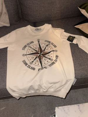Vit hoodie från Stone Island  - Vit hoodie från Stone Island med stor kompass- och logotypprint på bröstet samt klassisk patch på ärmen. Mjuk bomullskänsla och ribbade muddar. Perfekt för dig som gillar streetwear och vill sticka ut med en ikonisk design. Passar storlek m 