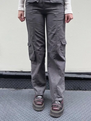 Brandy Melville lågmidjade grå cargobyxor - Säljer ett par grå cargobyxor med raka ben och stora fickor på sidorna. Byxorna är egentligen i bättre skick än vad de ser ut jag äger bara inget strykjärn tyvärr. 