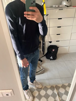 Mörkblå stickad tröja från J Lindeberg - Snygg mörkblå stickad tröja från J Lindberg med diskret strukturmönster. Tröjan har rund halsringning, långa ärmar och ribbade muddar vid ärmslut och nederkant. Perfekt att matcha med jeans för en clean look.