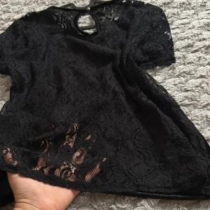 Svart spets tröja med kort ärm - Säljer en svart topp i spets med rund halsringning och nyckelhålsöppning fram. Toppen har korta ärmar och ett transparent, blommigt spetsmönster som ger en elegant look. Perfekt för dig som vill sticka ut lite extra.