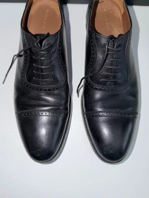 Svarta läderskor från Zara Man - Säljer ett par klassiska svarta läderskor från Zara Man. Skorna har rundad tå, snörning och diskreta broguedetaljer vid tåkappan. Insidan är fodrad i brunt och de har en stilren, tidlös design som passar till många olika looks.