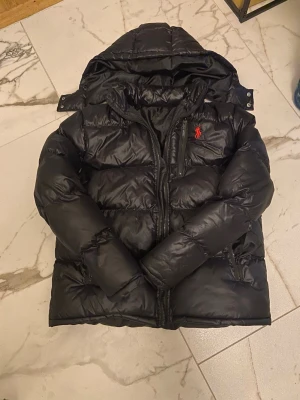 Svart pufferjacka från Ralph Lauren - Snygg svart pufferjacka från Ralph Lauren med huva och dragkedja framtill. Jackan har två sidofickor och den klassiska röda loggan broderad på bröstet. Perfekt för kalla dagar och riktigt skön att bära.