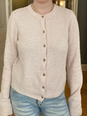 Ljusrosa kofta med knappar - Mysig ljusrosa kofta med rund halsringning och långa ärmar. Koftan har ribbade muddar och knäpps med små guldfärgade knappar framtill. Perfekt att slänga över en t-shirt eller linne för en enkel och stilren look.