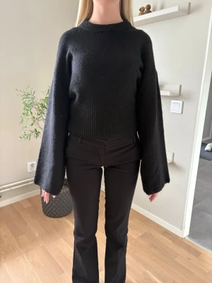 Svart tröja med vida ärmar - Säljer en svart stickad tröja med vida, långa ärmar och ribbad midja. Tröjan har en hög halsringning och är i mjukt material, perfekt för dig som gillar en stilren och trendig look.