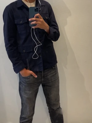 Mörkblå utilityjacka med knappar - Köpt från johnells knappast använt den säljer den för den här blivit för liten för mig. Snygg mörkblå utilityjacka med fyra framfickor och knäppning framtill. Jackan har en rak passform och hög krage, perfekt för dig som gillar en stilren och funktionell look. Tillverkad i bomull för en bekväm känsla. 