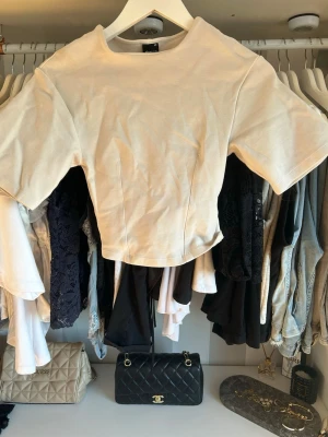 Beige croppad topp från Gina  - Trendig beige croppad topp från Gina med rund halsringning och korta ärmar. Tillverkad i mjuk bomull med en enkel, stilren design och markerade sömmar framtill. Perfekt att matcha med höga jeans eller kjol.