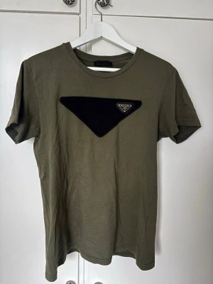 Olivgrön Prada t-shirt med logga - Snygg olivgrön t-shirt från Prada med svart triangulär logga på bröstet och liten röd Prada-detalj bak. Storlek M Nypris 6000kr!