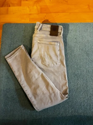 Ljusgråa Replay Ambass  jeans - Storlek 29. Säljer ett par ljusgråa Ambass jeans med klassisk femficksdesign. Jeansen har smal passform och raka ben, perfekta för en clean och modern look. Materialet är mjukt och stretchigt för extra komfort. Nypris: 1500kr, legat väldigt länge, varav det låga priset