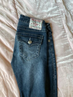 True Religion jeans rhinstones  - Säljer ett par mörkblå jeans från True Religion med bootcut och ikoniska rhinstones, stenar på sidorna. Jeansen har en snygg ficka med knapp bak samt en vit logga för true religion. Dem är uppsydda hos en skräddare skriv för mått! Snygga jeans som passar till allt, de är de lilla extra. Pris går att diskutera skriv för frågor!! 💓💓😇😇