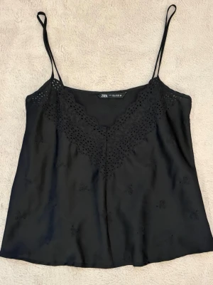 Svart linnetopp med spets från Zara - Snygg svart linnetopp från Zara med tunna axelband och v-ringad spetskant framtill. Tillverkad i ett glansigt material som ger en lyxig känsla. Perfekt att styla med jeans eller kjol för en trendig look.