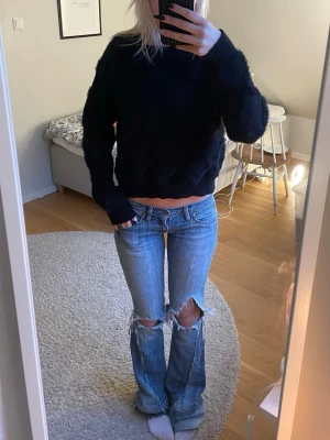 Svart fluffig stickad tröja - Mysig svart stickad tröja med fluffig textur och rund hals. Tröjan har långa ärmar och en avslappnad passform som passar perfekt till jeans eller kjol. Materialet ser ut att vara en mjuk blandning, troligen akryl eller polyester.