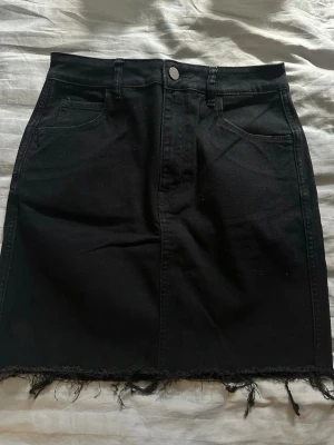 Svart jeanskjol från HM - Snygg svart jeanskjol från HM i klassisk femficksmodell. Kjolen har rå kant nertill för en trendig look och stängs med knapp och dragkedja framtill. Perfekt till vardags med en t-shirt eller stickad tröja.