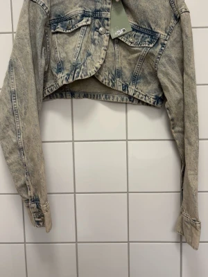 Cropped ljusblå jeansjacka - Säljer en trendig cropped jeansjacka i ljusblå tvätt med slitna detaljer. Jackan har två bröstfickor med knappar och klassisk krage. Perfekt för dig som vill ha en cool och avslappnad look. Lång ärm och snygg passform.