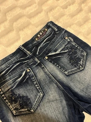 Mörkblå jeans med broderade fickor - Snygga mörkblå jeans från EDI Fashion Jeans med raka ben och slitningar. Bakfickorna har detaljerade blommiga broderier och små nitar. 🪽
