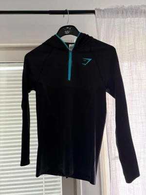 Svart hoodie från Gymshark - Säljer en svart långärmad hoodie från Gymshark med turkos dragkedja och logga på bröstet. Tröjan är i ett stretchigt funktionsmaterial som passar perfekt till träning eller löpning. Snygg och sportig design med åtsittande passform.