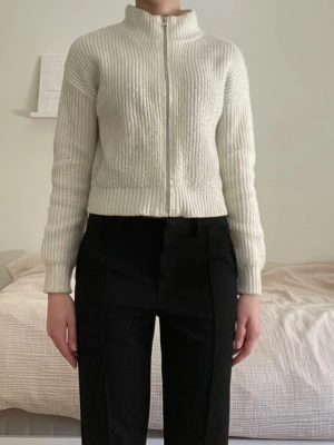 Vit ribbad ziptröja med hög krage - Säljer en stilren vit ribbstickad tröja med dragkedja framtill och hög krage. Tröjan är något croppad och har långa ärmar. Perfekt för dig som gillar minimalistisk och clean stil. Den har en liten brun fläck och skiftar lite i grått på vissa ställen på grund av användning.