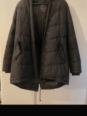 Svart dunjacka från Everest - Säljer en svart dunjacka från Everest med stor huva och dragkedja. Jackan har en långärmad design och är perfekt för kalla dagar. Den har praktiska fickor och en stilren look. Köptes från Stadium för några år sedan. Som ses på bilden är ena dragskon borta, men det går att fixa!