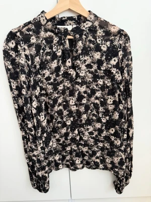 Inwear  - Blommig svart och beige blus - Trendig blus med svart och beige blommönster, rund halsringning och liten knytning fram. Långärmad modell med lätt puff vid axlarna och smala muddar vid ärmslut. Perfekt för dig som gillar en stilren men ändå lekfull look.