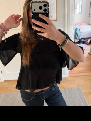 Svart transparent volangblus - Säljer en svart blus med volanger och transparent tyg. Blusen har korta, vida ärmar och en lös passform som ger en luftig känsla. Perfekt att styla med jeans eller kjol för en trendig look. Märke och material är ej angivet.