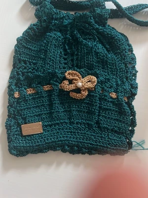 Handvirkad grön ryggsäck med blomdetalj - Unik handvirkad ryggsäck i mörkgrönt garn med guldfärgade detaljer och en virkad blomma med pärla framtill. Väskan har dragskostängning och axelband i samma material. Perfekt för dig som gillar boho-stil och vill sticka ut.