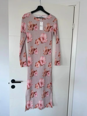 Lång blommig klänning från BYIC - Långärmad klänning från BYIC i en ljus beige ton med stora rosa och bruna blommor. Klänningen har en figurnära passform och är tillverkad i ett stretchigt, ribbat material. Perfekt för dig som gillar romantiska och feminina plagg.