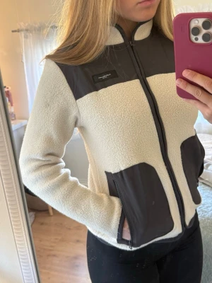 Vit fleecejacka Peak Performance - Säljer en vit fleecejacka från Peak Performance med svarta detaljer på axlar och fickor. Jackan har hel dragkedja framtill, höga fickor med dragkedja och stårkrage. Perfekt för lager-på-lager och riktigt skön att ha på sig. Liten fläck på vänster arm/ axel men den går nog att tvätta bort.