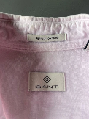 Ljusrosa oxfordskjorta från Gant - Säljer en klassisk ljusrosa oxfordskjorta från Gant med bröstficka och diskret broderad logga. Skjortan är tillverkad i bomull och har en stilren look som passar perfekt till många olika outfits.