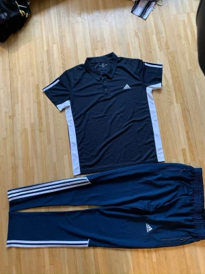 Adidas svart träningsset med vita detaljer - Säljer ett svart träningsset från Adidas med vita ränder och detaljer. Setet består av en pikétröja med krage och knappar samt ett par matchande träningsbyxor med klassiska Adidas-ränder längs benen. Perfekt för träning eller chill. Materialet är mjukt och andas.
