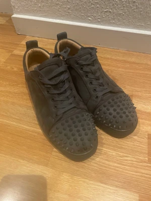 Gråa Louboutins  sneakers med nitar - Unika gråa sneakers med mattsvarta nitar på tån och ovandelen. Skorna har snörning, låg profil och är tillverkade i mockaliknande material. Insidan är ljusbeige och sulan är svart. Perfekta för dig som vill sticka ut med din stil. Några nitar fattas se sista bild men syns knappt. Superfina annars. Passar 43-44