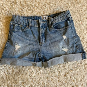 Blå jeansshorts med slitningar - Säljer ett par blå jeanshorts från lager 157 med slitna detaljer framtill och uppvikta benslut. Klassisk femficksmodell med knapp och dragkedja. Perfekta för varma dagar och enkel att matcha med olika toppar.