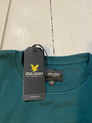 Mörkgrön sweatshirt från Lyle & Scott - Mörkgrön oversized sweatshirt från Lyle & Scott Junior med rund halsringning och en stor ficka med lock på bröstet. Tröjan har långa ärmar och ribbade muddar vid ärmslut och nederkant. Perfekt för en avslappnad och trendig stil.