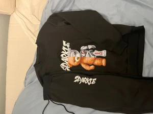 Svart set med björntryck från Sparkle - Säljer ett svart set från Sparkle med sweatshirt och byxor. Tröjan har ett stort tryck av en björn i rymddräkt och texten 'Sparkle' i vitt. Byxorna har matchande texttryck och dragsko i midjan. Perfekt för en avslappnad och cool stil.