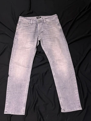Grå tvättade jeans Tapered Mike - Snygga grå jeans med tvättad look från Tapered Mike. Modellen har raka ben och klassisk femficksdesign. Jeansen har en ljusgrå ton med subtila slitningar och är tillverkade i ett mjukt denimtyg som ger en avslappnad vibe.