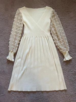 Vit omlottklänning med volangärm - Supersöt vit omlottklänning med ribbad struktur och transparenta långärmar i mesh med prickiga detaljer och volang vid ärmslut. Nedre delen är lätt utställd och överdelen har en v-ringad halsringning. Perfekt för dig som gillar romantisk och trendig stil.