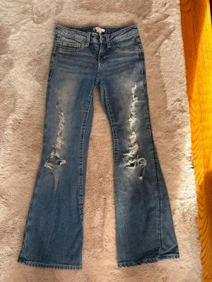 Blå bootcut jeans med slitningar - Säljer ett par blå bootcut jeans från Gina tricot med snygga slitningar och hål på framsidan. Jeansen har klassisk femficksdesign, dekorativa sömmar och knappar på bakfickorna. Perfekta för dig som gillar en avslappnad och trendig stil.