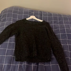 Mörkgrå stickad tröja - Mysig mörkgrå stickad tröja med rund halsringning och långa ärmar. Tröjan har en något lös passform och ribbstickade muddar vid ärmslut och nederkant. Perfekt för kyliga dagar och enkel att matcha med olika outfits.