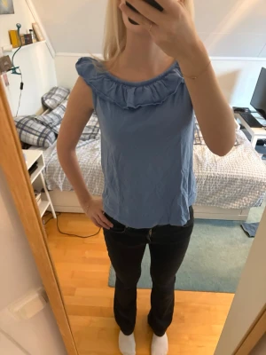 Blå ärmlös topp med volang - Söt blå ärmlös topp med volangdetalj runt halsringningen. Toppen har en avslappnad passform och är perfekt för varma dagar. Tillverkad i ett mjukt bomullsliknande material och har en enkel, stilren look.