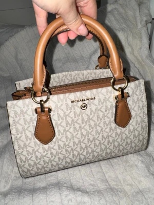 Michael Kors Marilyn hansväska , helt ny  - Snygg beige handväska från Michael Kors med klassiskt MK-mönster och detaljer i brunt läder. Väskan har två robusta handtag, guldfärgade metalldetaljer och dragkedja upptill. Perfekt för dig som gillar stilrena accessoarer med lyxig känsla.
