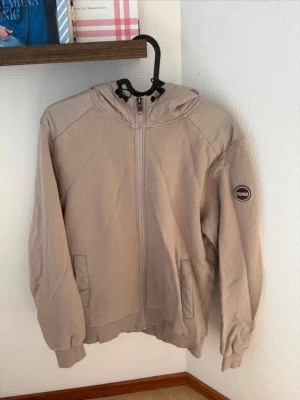 Beige hoodie från Colmar med dragkedja - Snygg beige hoodie från Colmar med dragkedja framtill och två fickor. Har en stor huva och ribbade muddar vid ärmar och nederkant. Märkeslogga på ena ärmen. 