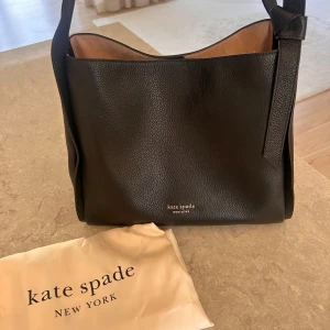 Svart handväska från Kate Spade - Stilren svart handväska från Kate Spade New York i mjukt, kornigt läder. Väskan har en enkel, fyrkantig form med bred axelrem och dekorativ knut på sidan. Insidan är beige och väskan stängs med magnetknapp. Perfekt för dig som gillar klassisk och tidlös design.