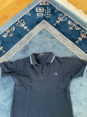 Mörkblå pikétröja från Fred Perry - Klassisk mörkblå pikétröja från Fred Perry med vitrandiga detaljer på krage och ärmslut. Tröjan har korta ärmar, knappslå framtill och den ikoniska Fred Perry-loggan broderad på bröstet. Perfekt för en stilren och sportig look.