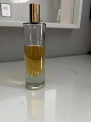 Zara Golden Decade Elixir parfym - Zara Golden Decade Elixir är en elegant parfym i en genomskinlig glasflaska med guldfärgat lock och spraymunstycke. Doften har en varm, gyllene ton och flaskan har en stilren, modern design som känns lyxig och trendig.