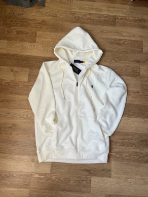 Vit hoodie från Polo Ralph Lauren - Snygg vit hoodie från Polo Ralph Lauren med dragkedja framtill och klassisk huva med dragsko. Ikoniska mörkblå loggan broderad på bröstet. Tillverkad i mjukt bomullsmaterial och har två fickor framtill. Perfekt för en clean och stilren look.