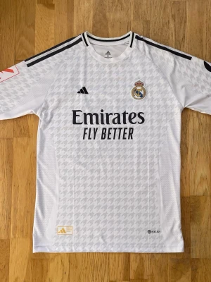 Real Madrid Jersey 24/25 ”Mbappé” - Säljer min helt nya och oanvända real Madrid träningströja från åren 24/25. På ryggen står det ”Mbappe(nr10)”. Inga defekter. Storlek medium. Axelbredd är 47cm och längden är 74cm! Material är jag osäker på men gissar på Nylon. Kom med frågor och bud. Skickas samma dag