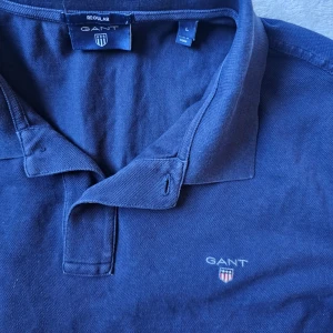 Marinblå pikétröja från Gant - Klassisk marinblå pikétröja från Gant med krage och knappar framtill. Tröjan har Gant-logga broderad på bröstet och är tillverkad i mjuk bomull. Perfekt till jeans eller shorts för en clean och stilren look.