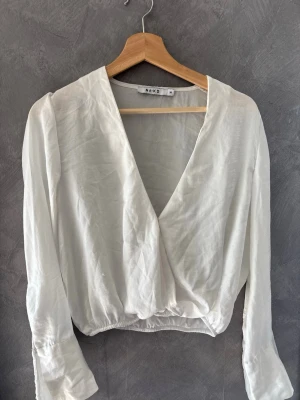 Vit omlottblus från NA-KD - Stilren vit omlottblus från NA-KD med djup v-ringning och långa ärmar. Blusen har en lätt croppad passform och är gjord i ett tunt, luftigt material som ger en avslappnad men chic look. Perfekt att matcha med höga jeans eller kjol.